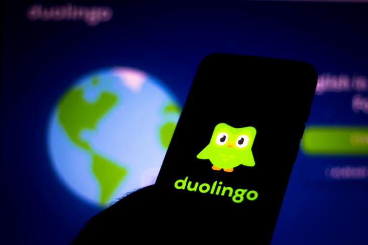 Esta imagem retrata o logotipo do aplicativo Duolingo em um smartphone com a página inicial do aplicativo aberta em uma tela ao fundo