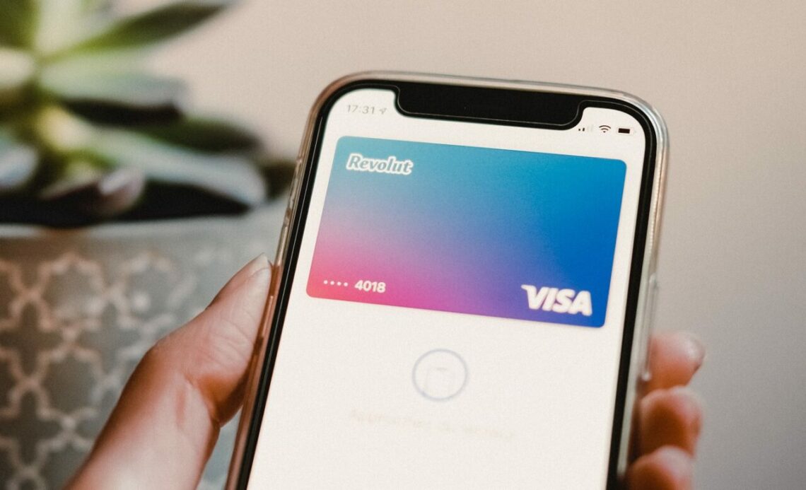 Uma pessoa segura um smartphone exibindo um cartão de pagamento virtual Revolut Visa na tela, com os últimos dígitos do número do cartão visíveis e um gradiente de cor no fundo.
