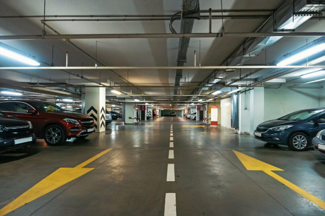 Interior de um estacionamento subterrâneo com carros estacionados em ambos os lados, faixas demarcadas e setas direcionais no chão. Pode haver um carregador para carros elétricos em segundo plano,