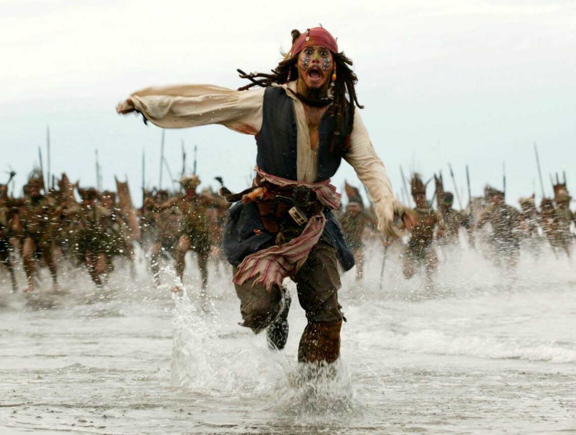 Johnny Depp, vestido de pirata, foge por águas rasas, claramente aterrorizado, com uma multidão de homens armados à distância, perseguindo-a.