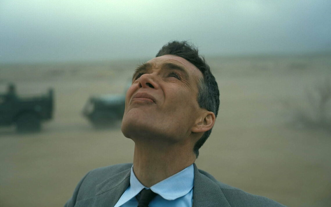 Um homem de terno e gravata olha para cima com uma expressão pensativa contra o pano de fundo de um deserto nebuloso com um veículo militar desfocado ao fundo.  Um still do filme Oppenheimer que será exibido no SkyShowtime.