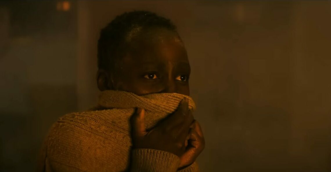 Quadro do filme Um Lugar Silencioso 3. Uma jovem de pele escura, vestindo um suéter cobrindo a boca e o nariz, parece assustada ou ansiosa, em uma sala com iluminação quente e escura.